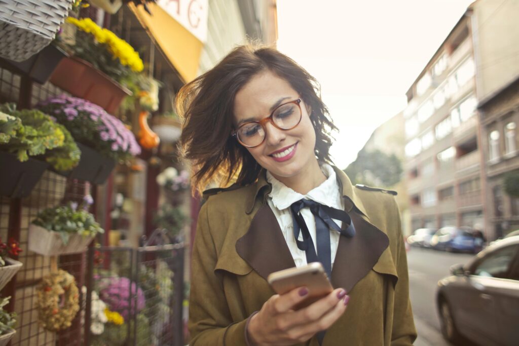 Fibra y Movil A smiling woman uses her phone outside a colorful urban flower shop on a sunny day.